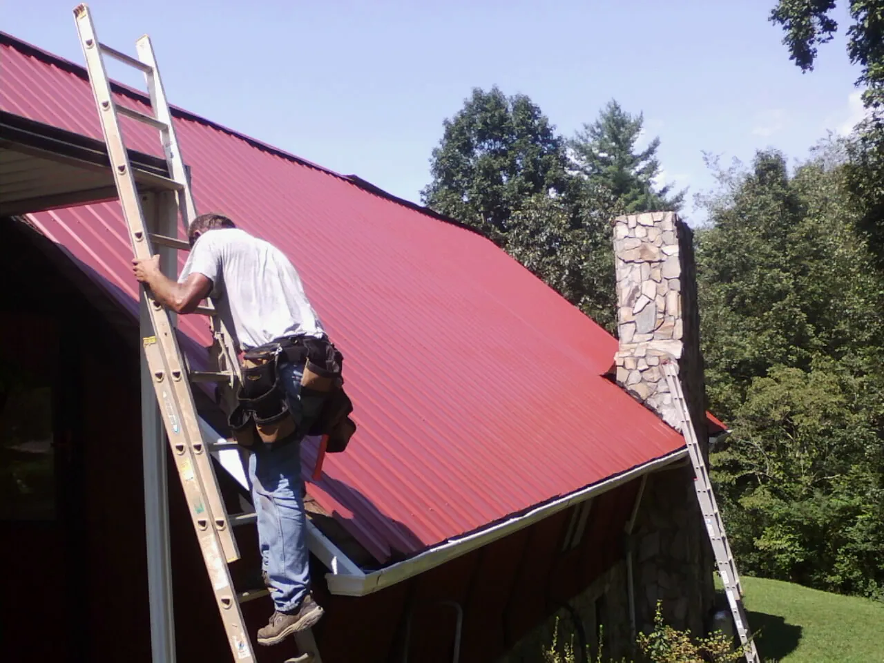 Roofer on a ladder beside a red metal roof and stone chimney in Ulm